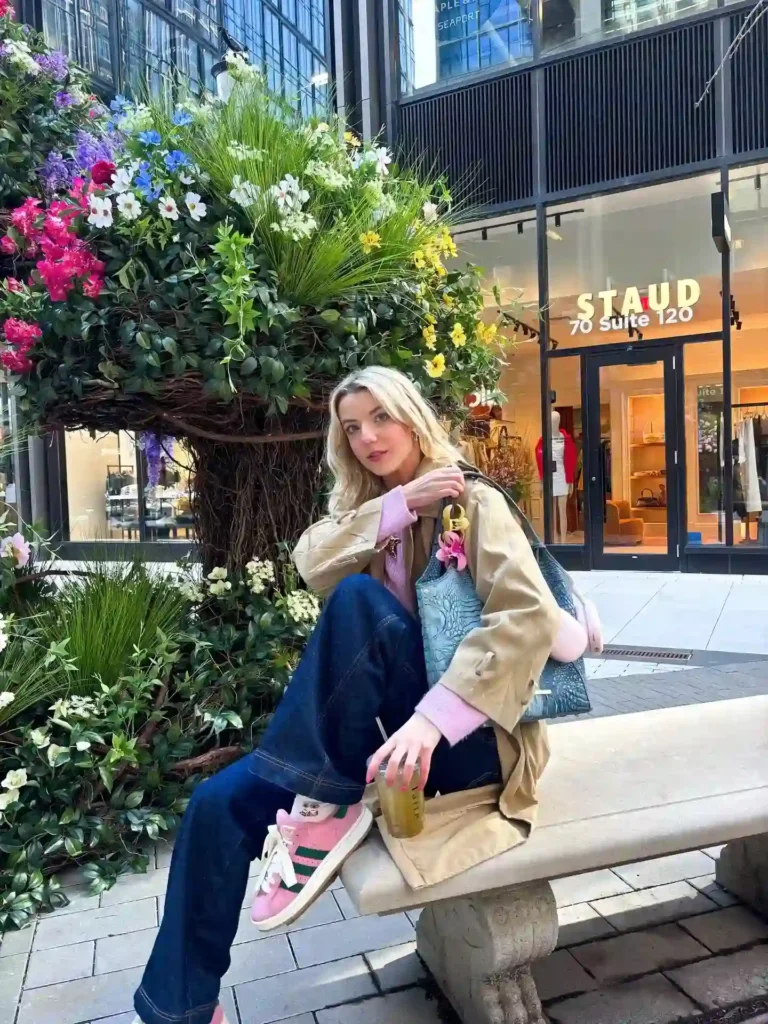 Stylish woman in casual attire sits among vibrant flowers, showcasing a trendy outfit near the STAUD store.