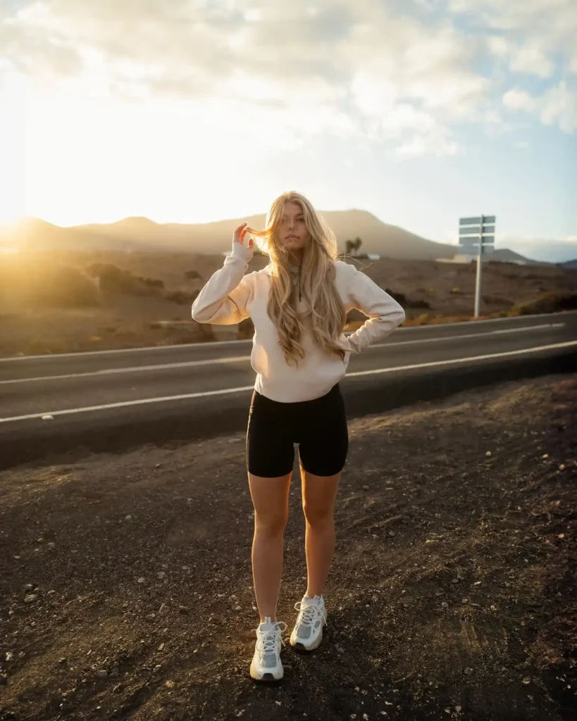Young woman in activewear poses by the roadside at sunset, showcasing a sporty style and relaxed vibe.