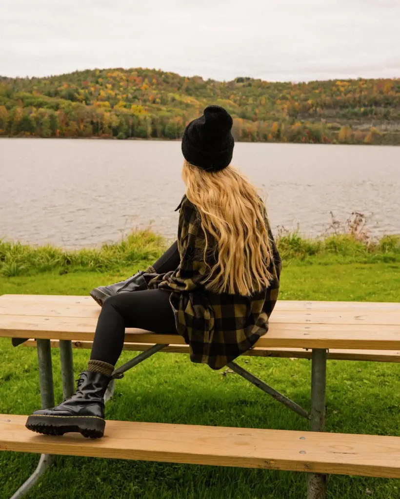 Woman sitting on a bench by a lake, wearing a black beanie and plaid shirt, surrounded by autumn foliage.