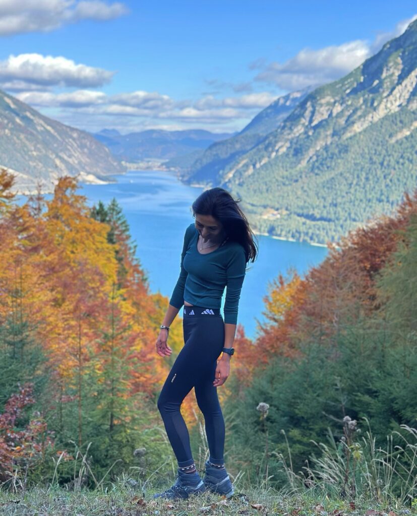 Young woman in athletic wear standing on a mountain overlooking a lake, surrounded by colorful autumn foliage and mountains.