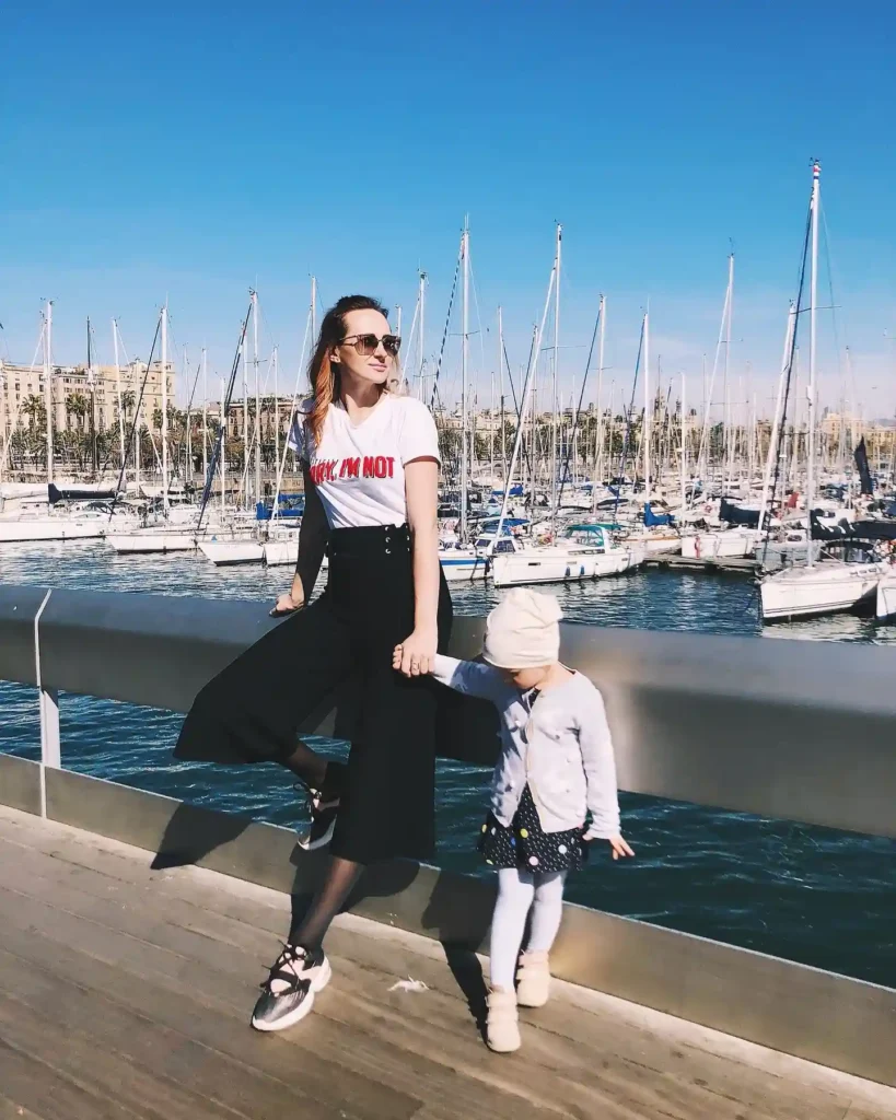 A woman in sunglasses poses by the marina with a child, enjoying a sunny day by the water and boats.