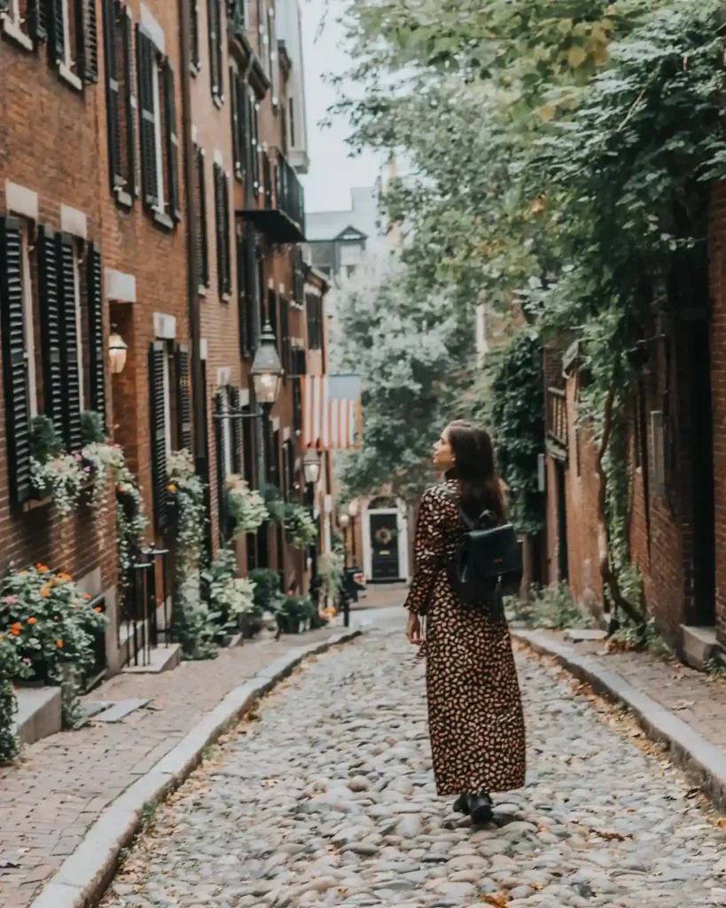 Woman in a patterned dress walking down a charming cobblestone street lined with historic brownstone buildings.