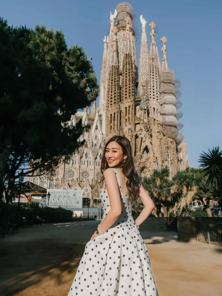 Young woman in a polka dot dress poses near Barcelona's Sagrada Familia under a clear blue sky.