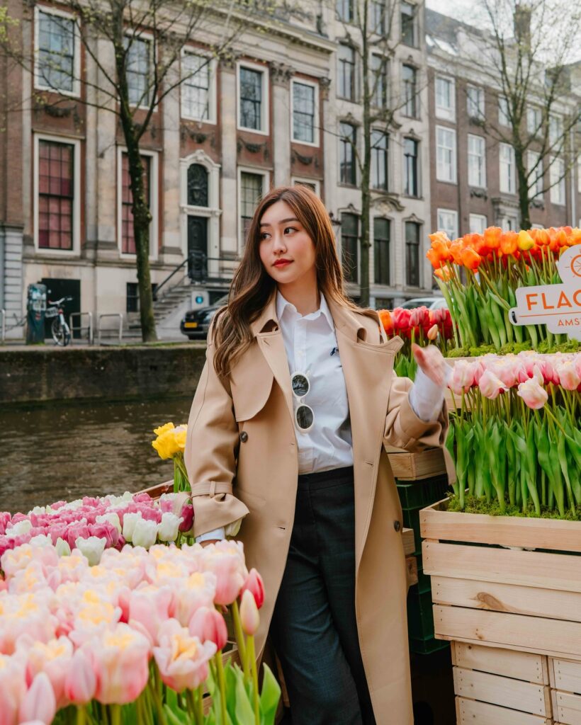 Woman in a trench coat stands beside vibrant tulip displays along a canal in Amsterdam, surrounded by historic architecture.