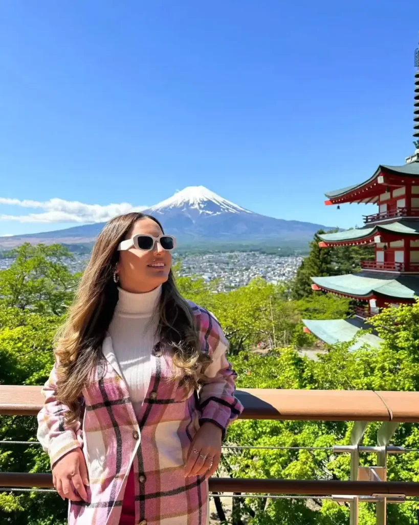 Young woman with sunglasses stands near a pagoda, enjoying a view of Mount Fuji on a sunny day.