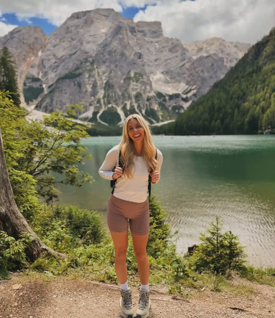 Smiling hiker with a backpack stands by a lake, surrounded by majestic mountains and greenery. Perfect for outdoor adventures.