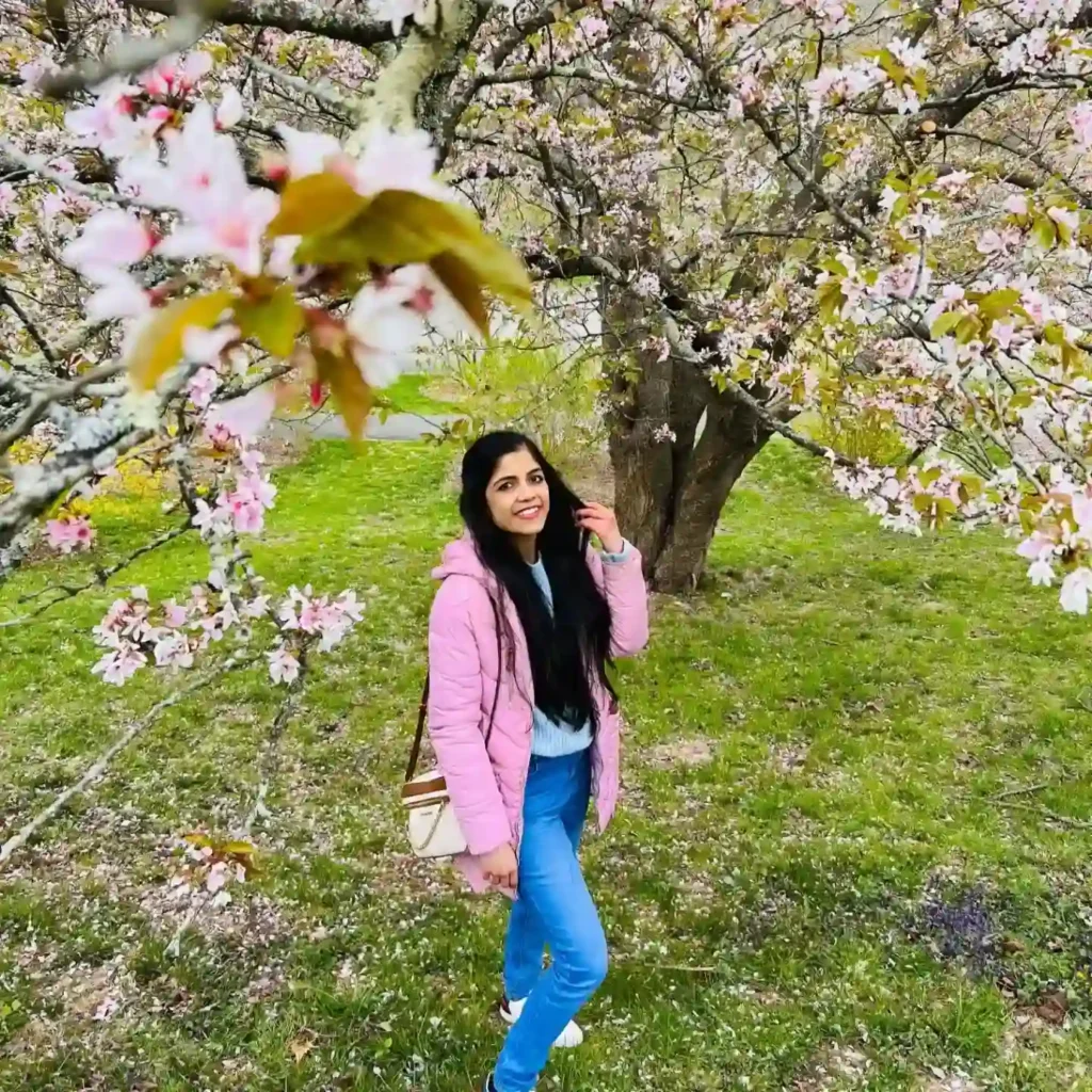 Young woman smiling in a pink jacket, standing under a blossoming tree with pink flowers in a green park.
