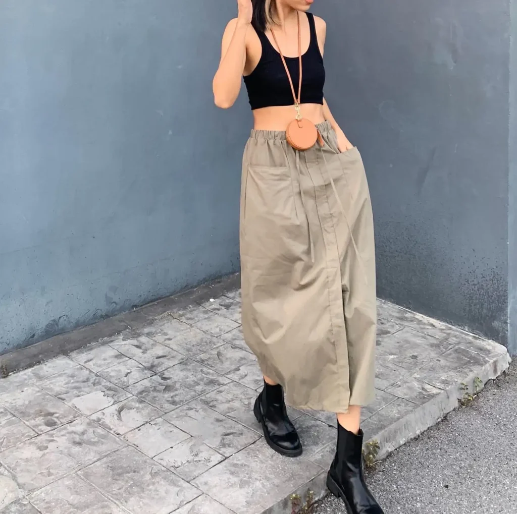 Woman in a black crop top and khaki maxi skirt, posing against a gray wall, showcasing a trendy urban outfit.