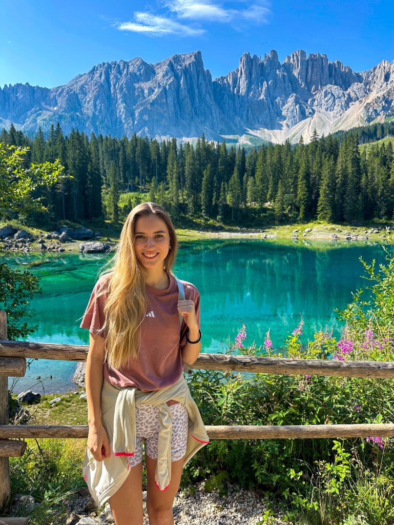 Young woman smiling by a stunning turquoise lake, surrounded by lush forests and towering mountains under a clear blue sky.