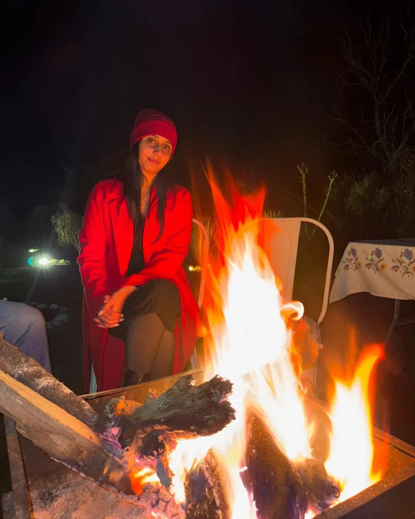 Young woman in a red coat and hat sitting by a campfire at night, enjoying the warmth and ambiance of the flames.