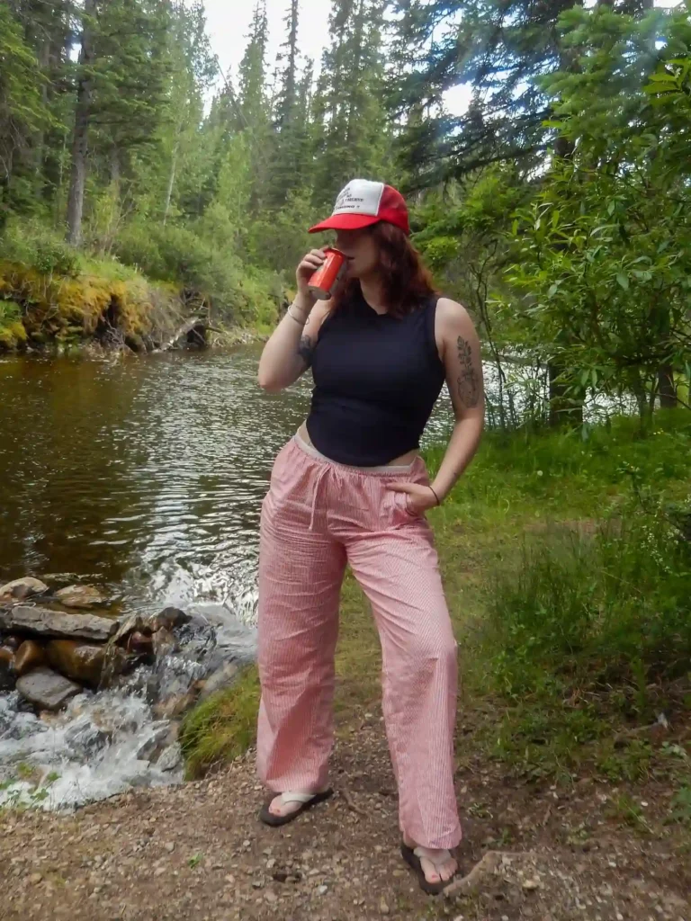 Woman sipping a drink by a serene river, surrounded by tall trees and lush greenery, in casual summer attire.