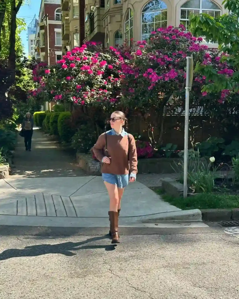 Young woman walking in a sunny urban area, surrounded by blooming pink flowers and greenery. Stylish casual outfit.