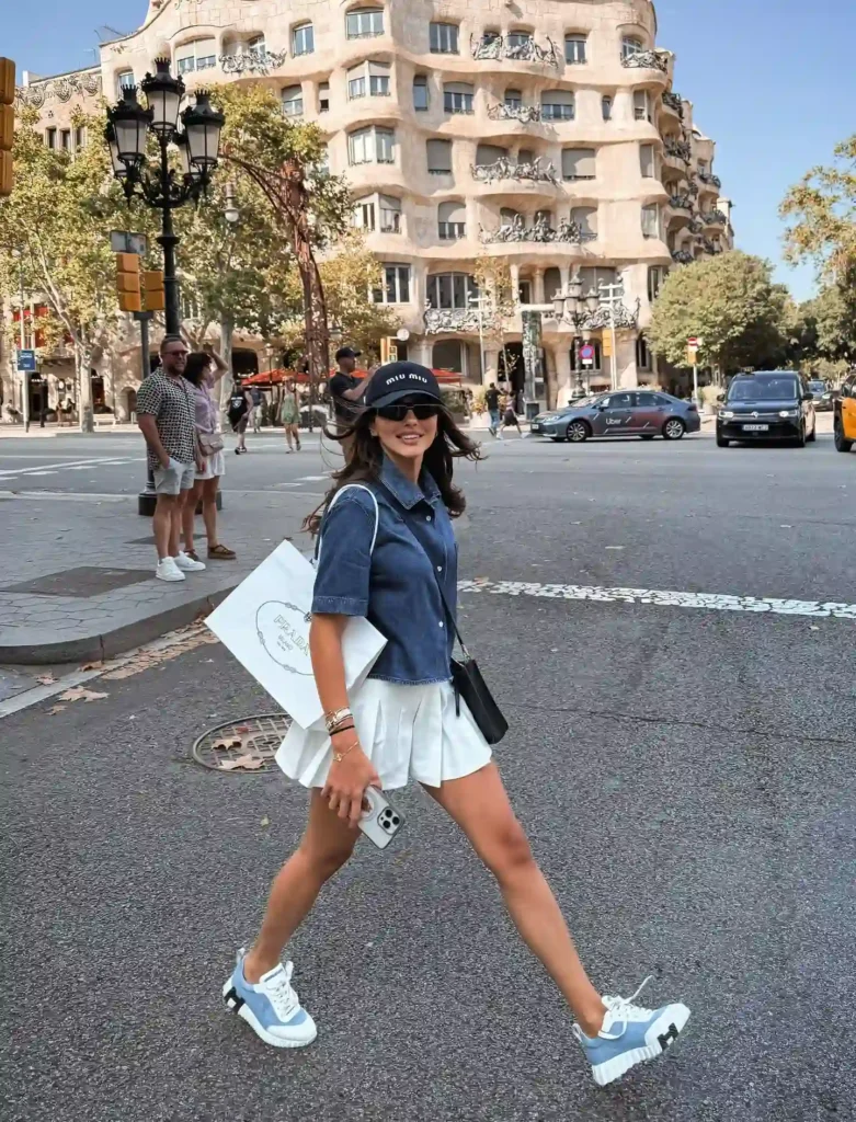 Stylish woman in a denim shirt and white skirt walking past iconic architecture and city life.