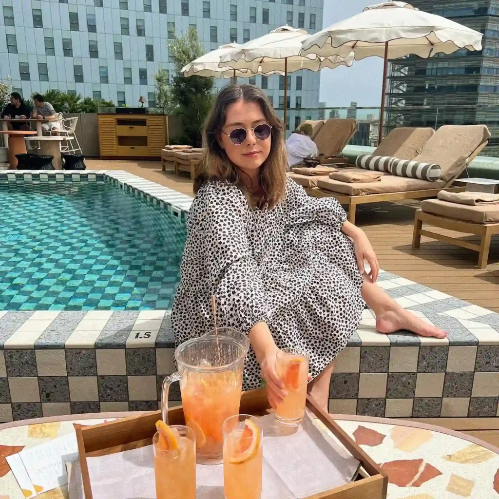 Woman in sunglasses relaxes by a poolside, enjoying refreshing drinks on a sunny day. Trendy attire and serene atmosphere.