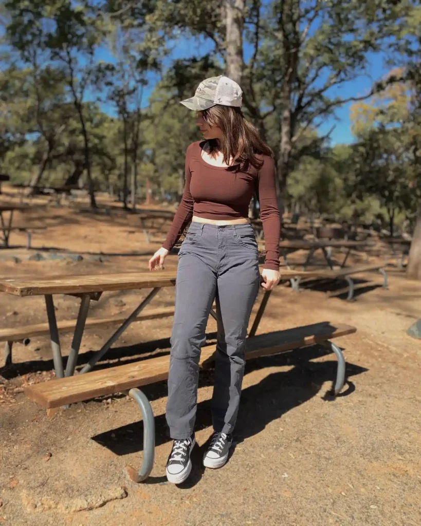 A woman wearing a brown crop top, gray pants, and sneakers stands outdoors near picnic tables under a clear blue sky.