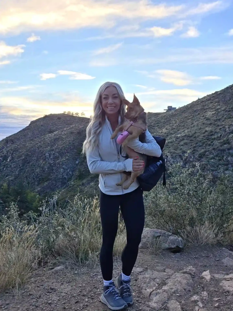 Smiling woman holding a dog on a hiking trail with mountains and a colorful sky in the background.