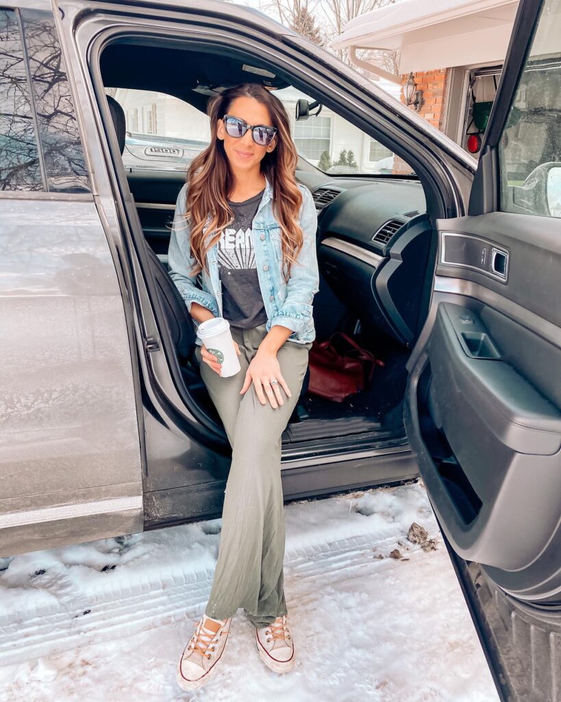 Woman in sunglasses and casual outfit sitting in a car doorway, holding a coffee cup amidst a snowy landscape.