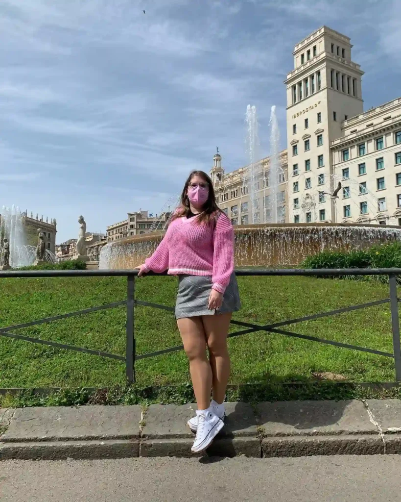 Young woman in a pink sweater and mask poses by a fountain in a vibrant city square. Sunny day with clear skies.