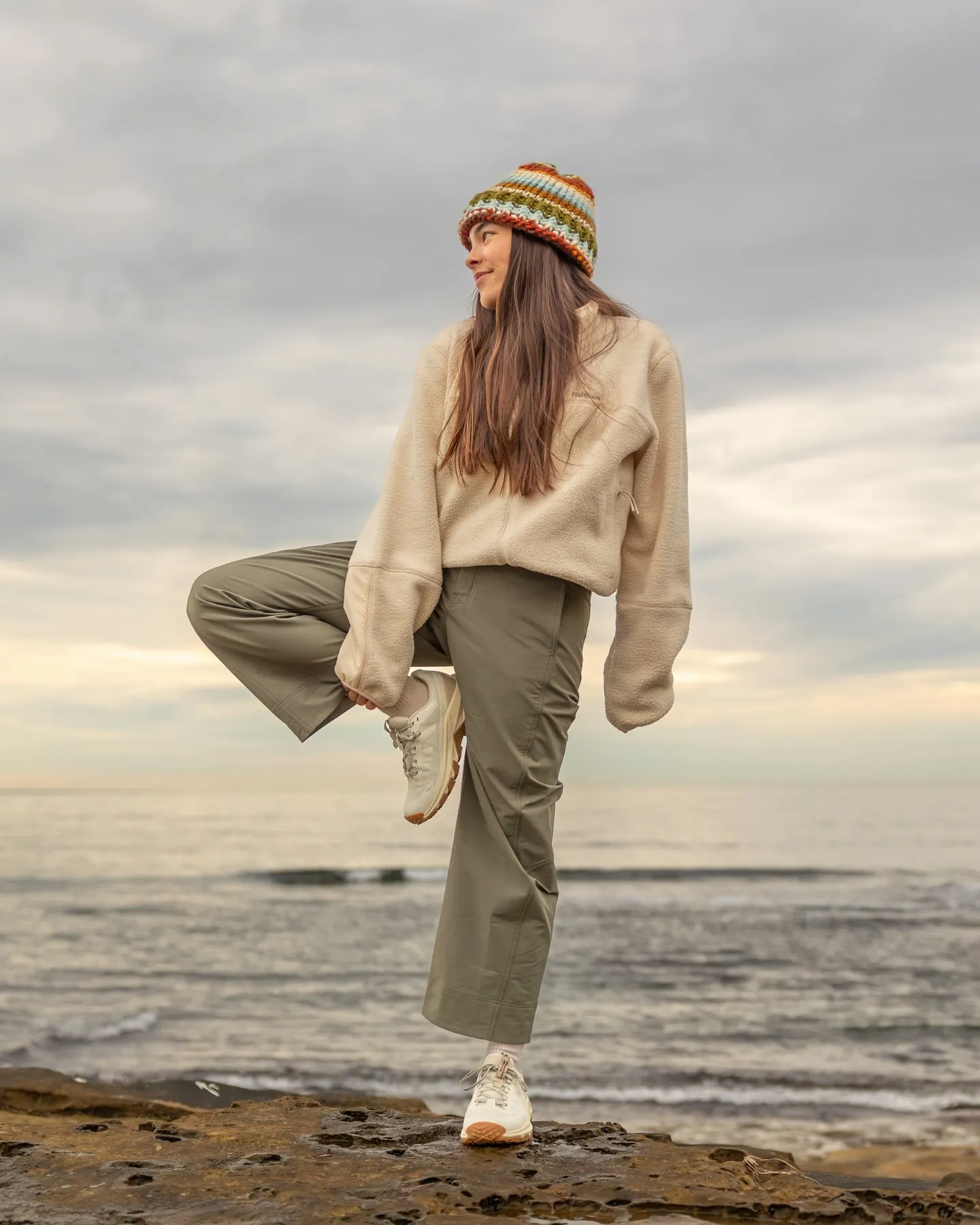 Young woman in cozy outfit and colorful hat poses by the ocean, showcasing casual style against a cloudy sky.