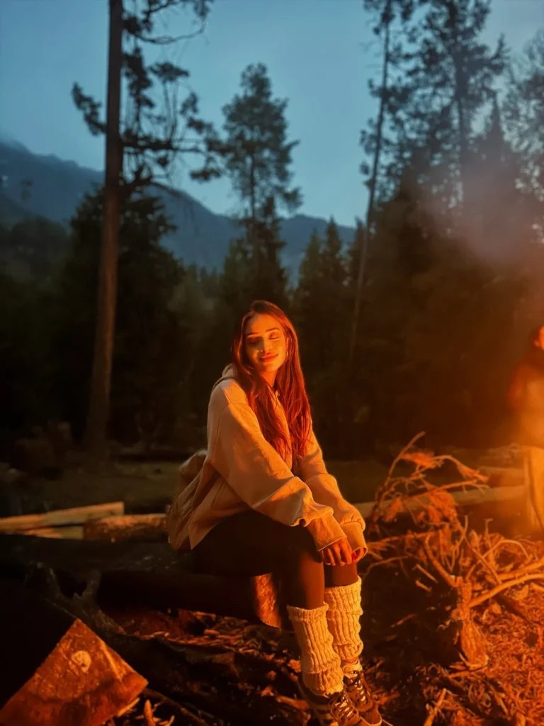 Young woman sitting by a campfire in the woods, enjoying a cozy evening with a peaceful mountain backdrop.