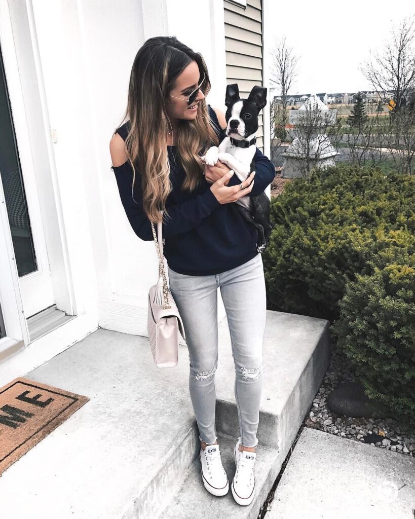 Young woman in sunglasses holds a black and white dog on her porch, wearing casual attire with a stylish handbag nearby.