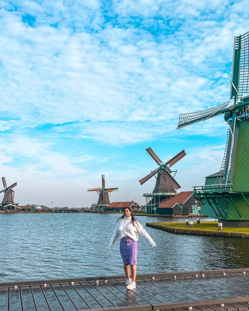 Woman standing by the water at Zaanse Schans, surrounded by traditional Dutch windmills and a vibrant blue sky.