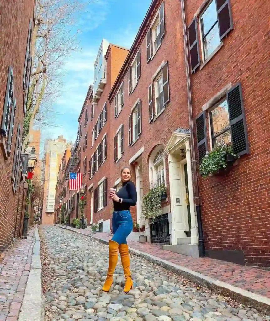 Young woman in stylish outfit poses on a cobblestone street, framed by historic brick buildings and an American flag.
