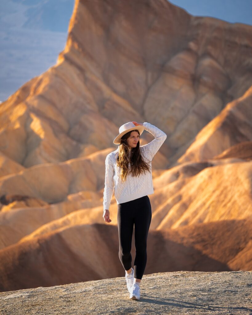 A woman in a white sweater and hat walking on a rocky landscape with colorful, textured hills in the background.