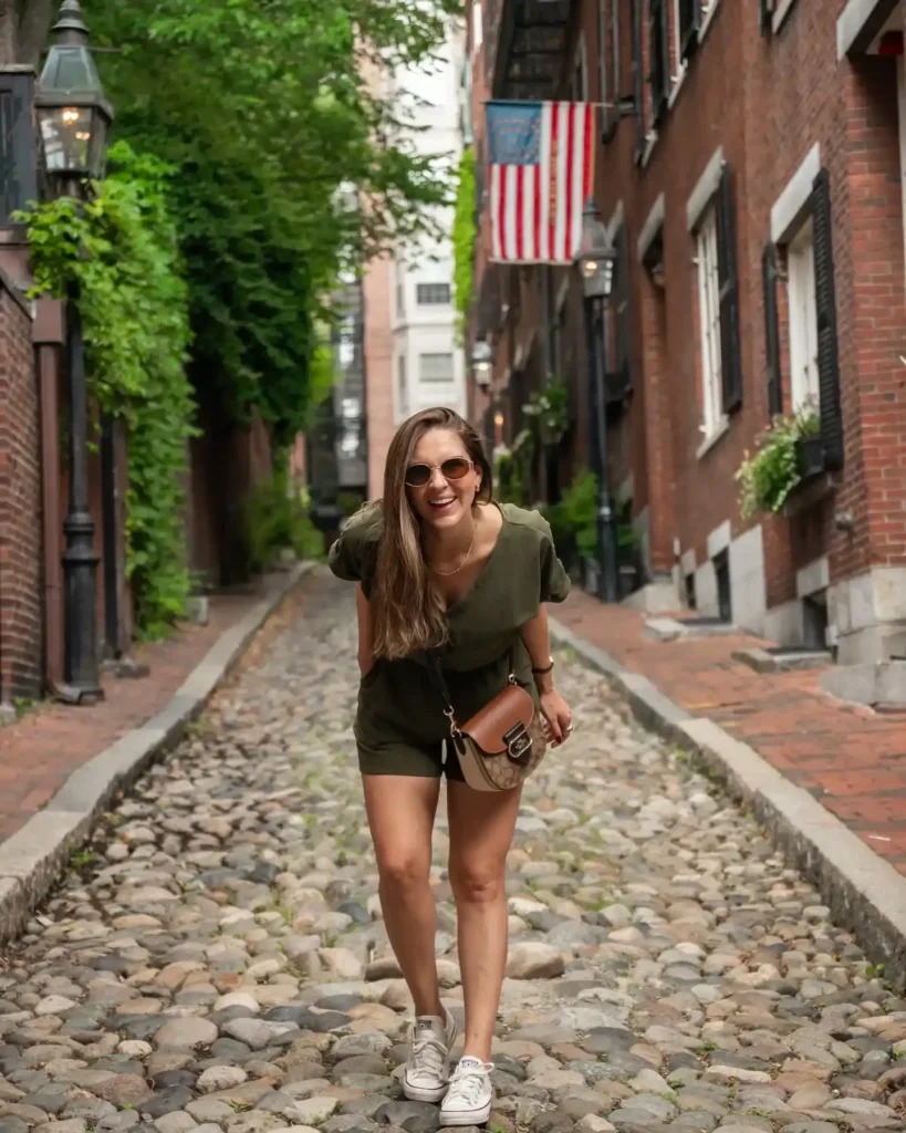 Smiling woman in sunglasses walking on cobblestone street, surrounded by lush greenery and historic brick buildings.