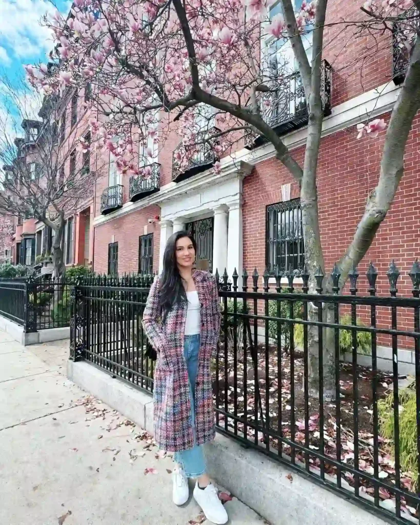Young woman in a stylish coat stands by a blooming magnolia tree in a picturesque urban setting.