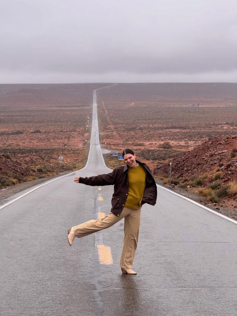 Woman joyfully poses on a long, empty road surrounded by scenic, rugged landscape under overcast skies.