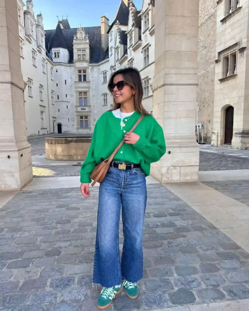 Fashionable woman in green sweater and wide jeans smiling in a historic courtyard with elegant architecture.