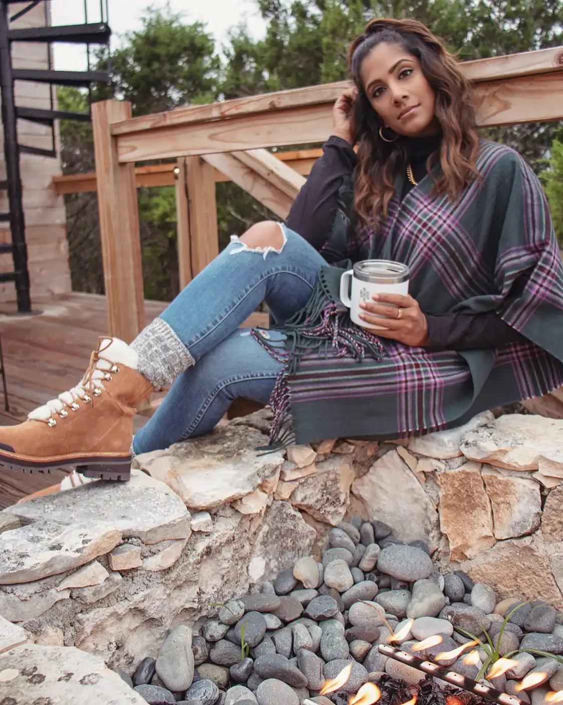 Woman in a stylish poncho sitting by a stone fire pit, sipping from a mug, wearing cozy jeans and rugged boots.
