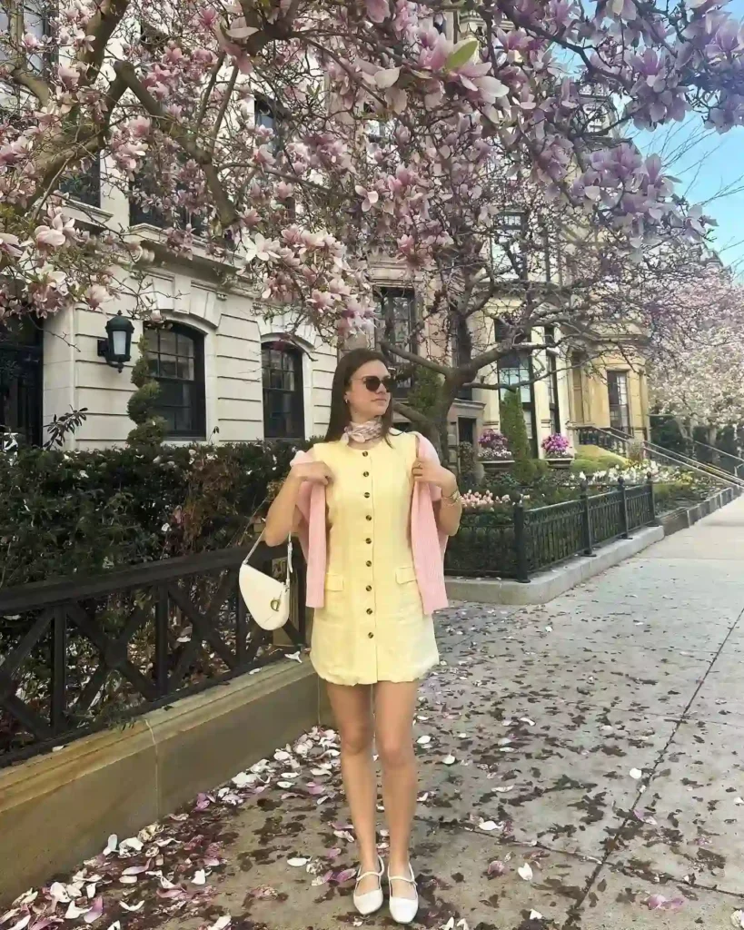 Young woman in a yellow dress and pink sweater stands under blooming magnolias on a sunny street, enjoying springtime.