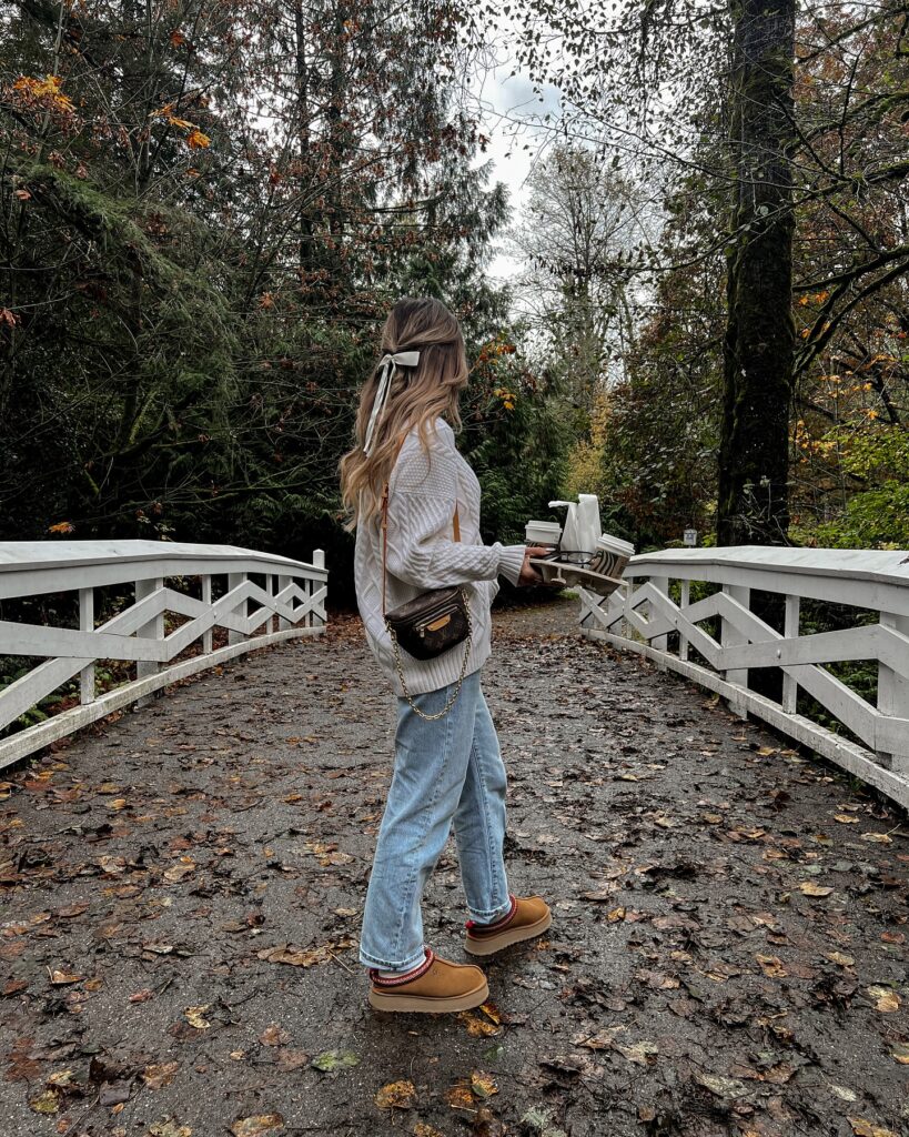 Woman in cozy sweater and jeans stands on a leaf-covered bridge, holding a book, surrounded by nature in autumn.