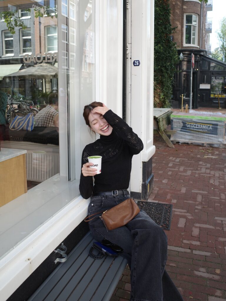 Young woman laughing while seated on a bench outside a café, holding a coffee cup in a stylish urban setting.