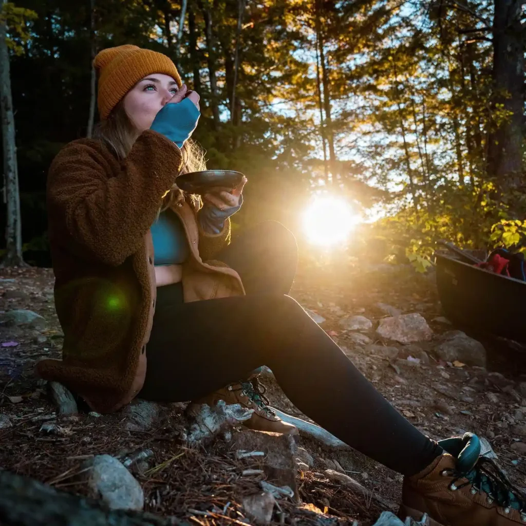 Woman enjoying a meal beside a canoe in a serene forest at sunset, wrapped in a cozy jacket and hat.