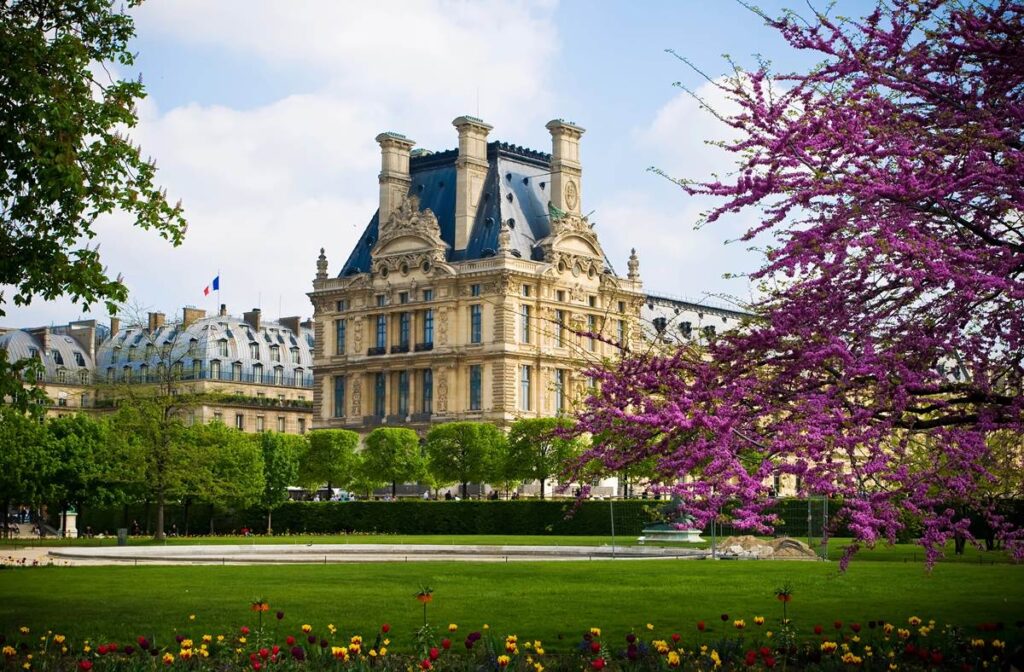 Historic Louvre Palace surrounded by blooming gardens and vibrant flowers on a sunny day in Paris.