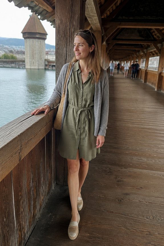 Woman in olive green dress and gray cardigan leaning on a wooden walkway railing along a lake, historic tower in background.