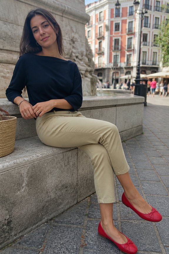 Woman seated on stone steps in a city square, wearing a black top, beige pants, and red flats; urban backdrop.