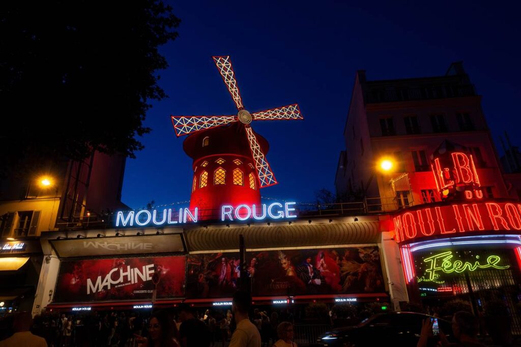 Iconic Moulin Rouge windmill illuminated at night in Paris, with vibrant neon lights and bustling street below.
