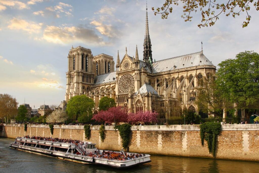 Gothic cathedral by river with blooming trees and a tour boat, under a partly cloudy sky.