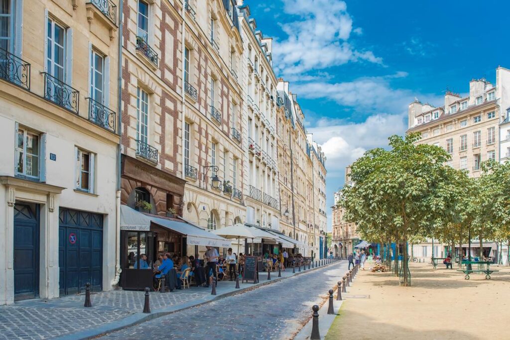 Charming Parisian street with outdoor cafes, historic buildings, and trees under a sunny sky.