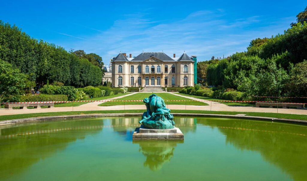 Elegant mansion with lush gardens and a central fountain sculpture under a clear blue sky.