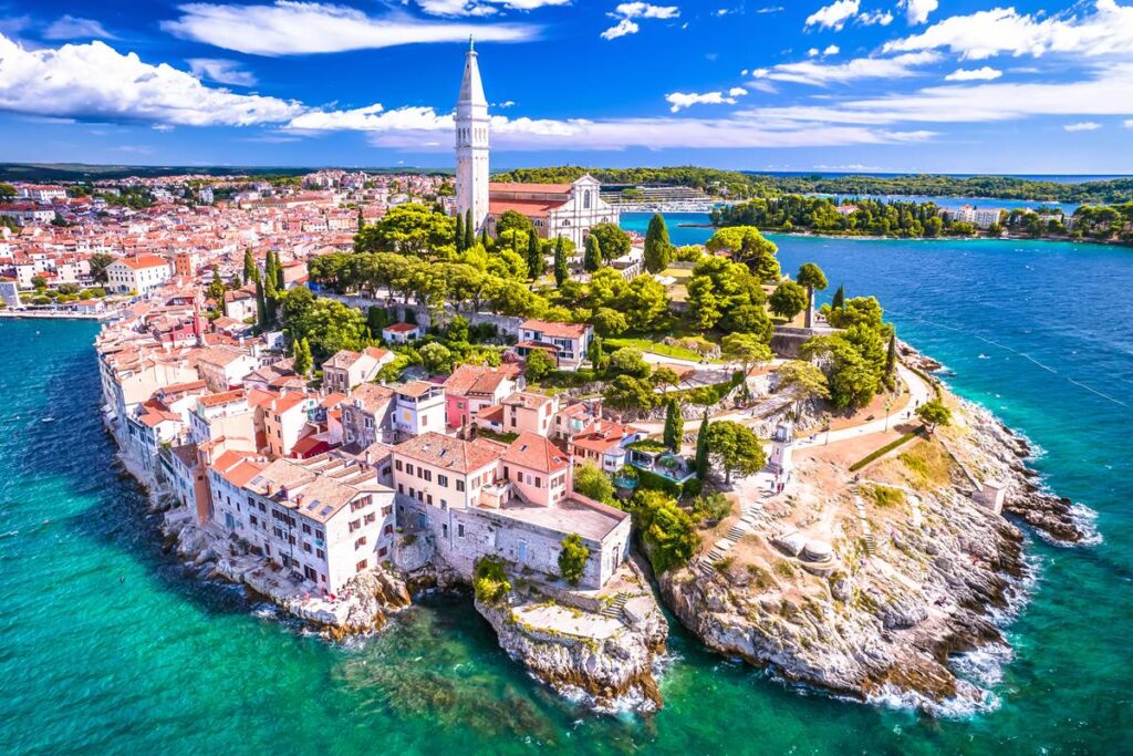 Aerial view of Rovinj, Croatia, featuring St. Euphemia's Church, ocean, and vibrant rooftops on a sunny day.