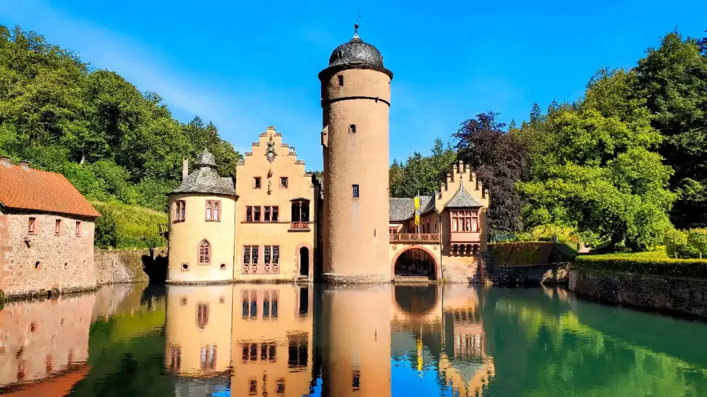 Charming castle reflected in peaceful water, surrounded by lush greenery and bright blue skies. Perfect for fairy-tale lovers.