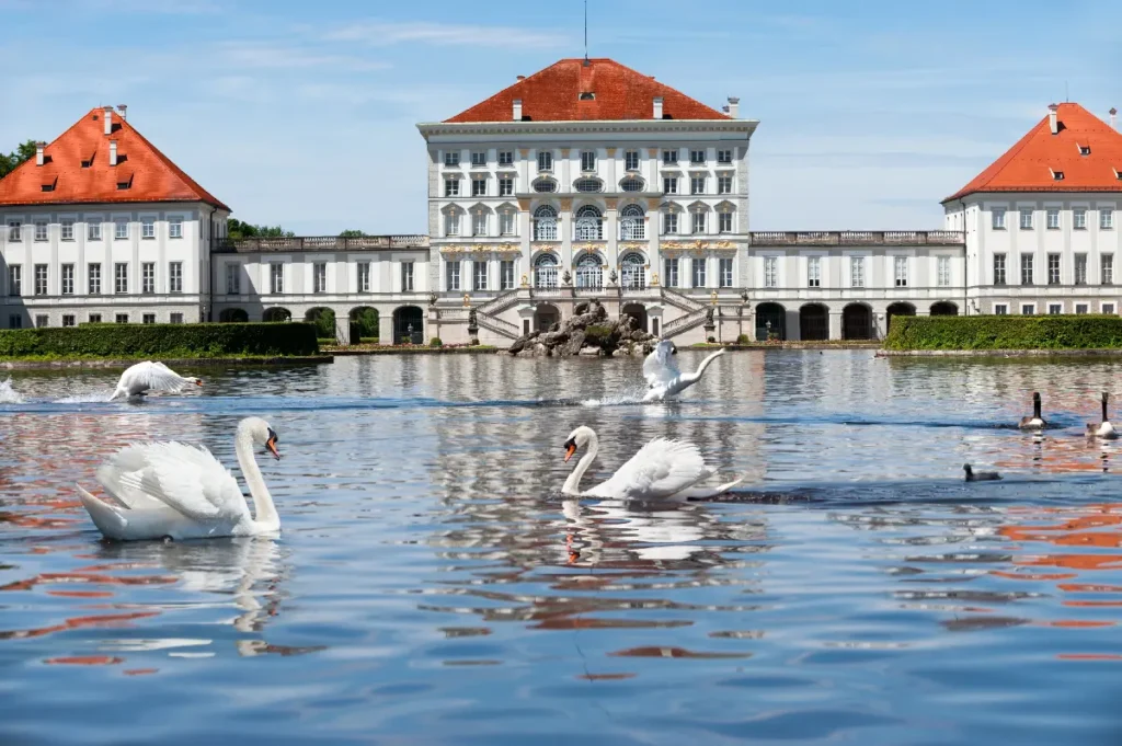 Beautiful palace with red-roofed architecture, surrounded by swans swimming in a calm lake on a sunny day.