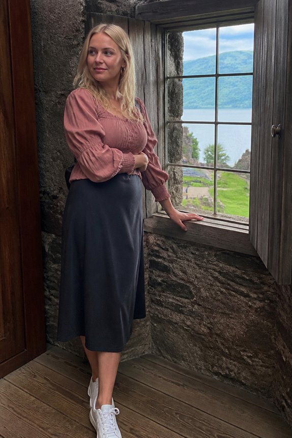 Blonde woman in a pink blouse and navy skirt stands beside a rustic stone window, overlooking a lake and mountains.