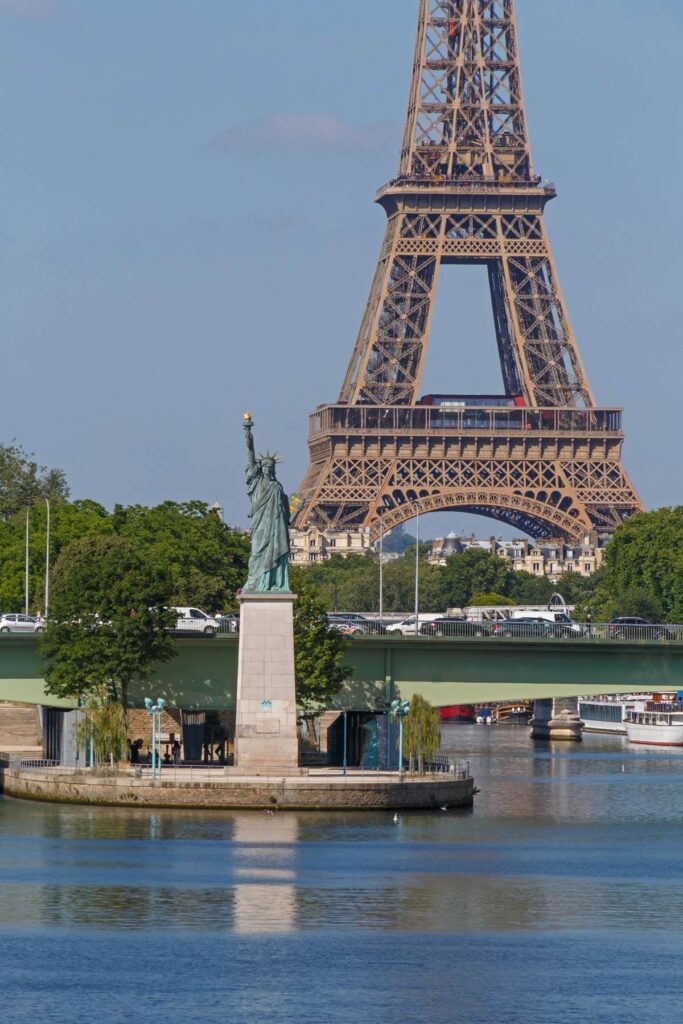 Mini Statue of Liberty and Eiffel Tower on a sunny day in Paris, with lush greenery and blue skies.