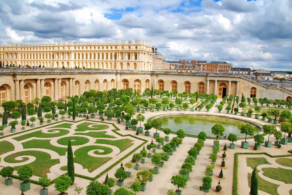 Ornate gardens with manicured hedges at a historic palace under a partly cloudy sky.